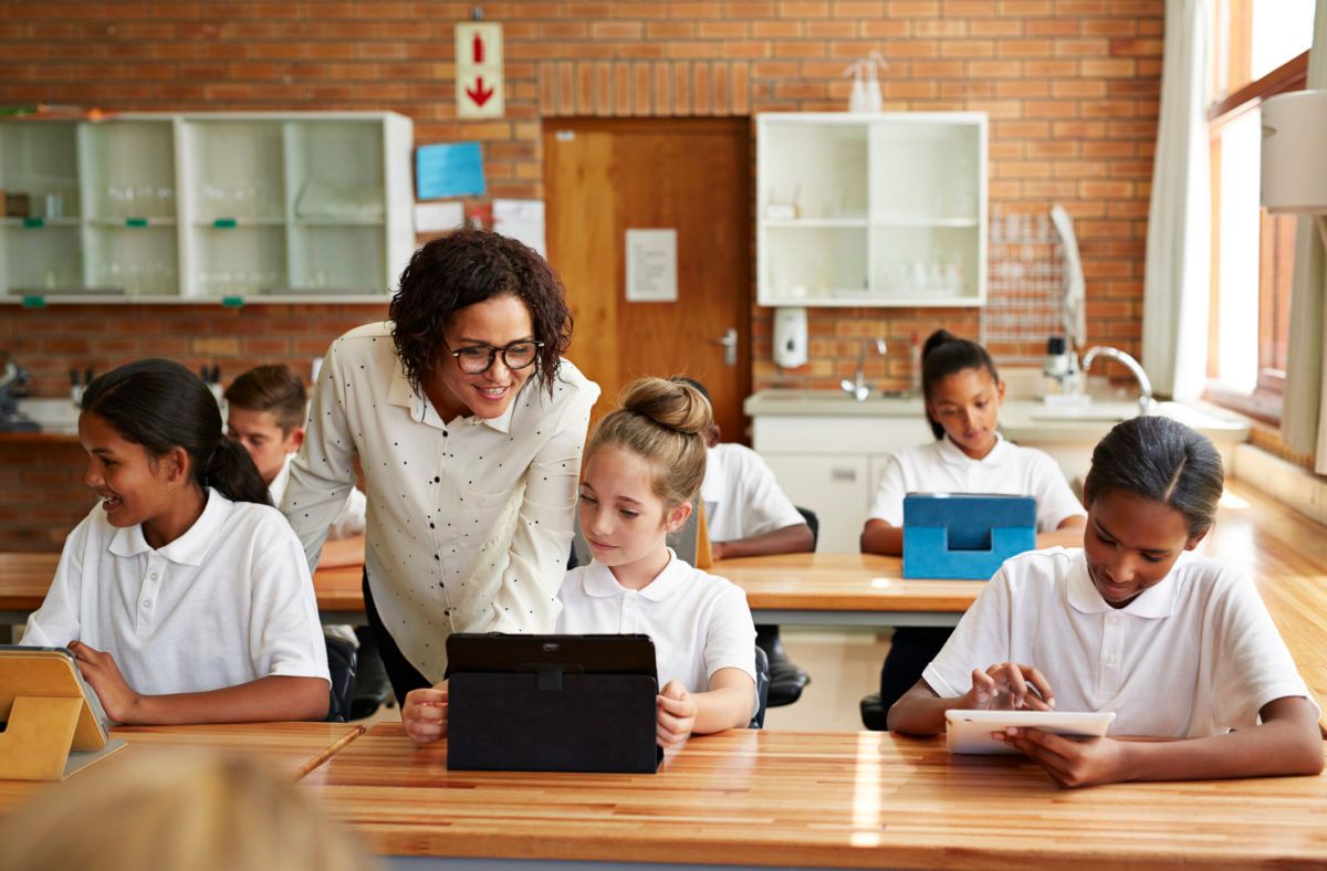 Teacher helping students at desks with tablet computer and papers in classroom