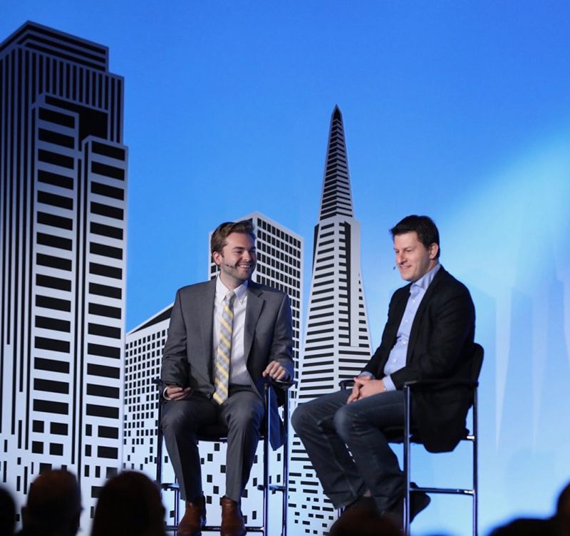 Two men in conversation on a stage with a San Francisco skyline backdrop