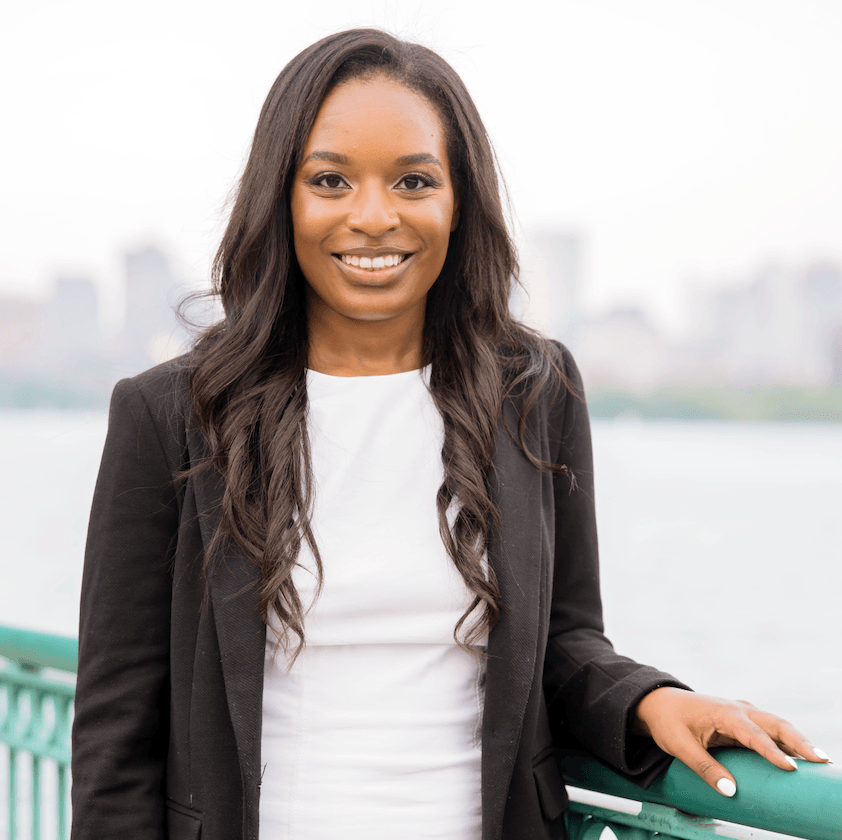 Photo of a Black woman with long straight hair in a white tee and black jacket, standing at the railing of a city waterway.